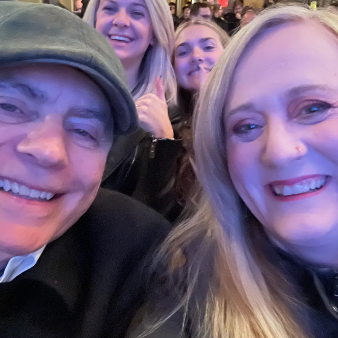Patric (a man wearing a green cap) and Christal (a women with long blonde hear) are smiling as they are photobombed in the background by two young women.
