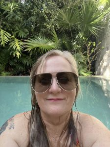 A woman in a pool in front of tropical plants. The woman is wearing large sunglasses and her blonde hair is wet. She has a tattoo barely visible on her right shoulder. She has a small smile. 